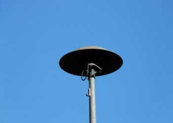 Masmavi açık gökyüzü önünde direğe monte edilmiş gri renkli yuvarlak siren sistemi.