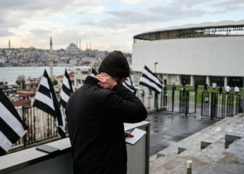 Siyah beyaz bayrakların asılı olduğu statta not alan futbol antrenörü