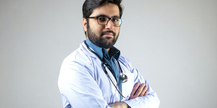 Kollarını bağlamış, gözlük takan doktor kameraya ciddi bir şekilde bakıyor.