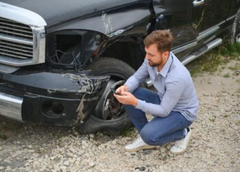 Hasar gören siyah bir aracın önünde çömelip telefona bakan adam.