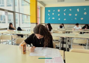Sınıfta sırada sınav olmaya çalışan gençler, önlerinde test kitapları ve içecek kutusu.