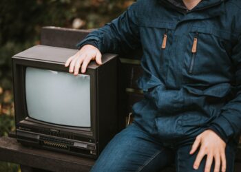 Mavi montlu bir adam, açık havadaki bankta duran eski bir televizyona dokunuyor.