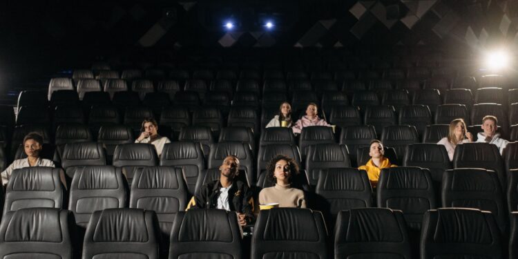 Karanlık bir sinema salonunda koltuklarında oturan ve ekrana bakan seyirciler.