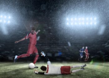 Futbol sahasında yağmur altında topa vuran kırmızı formalı futbolcu.