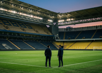 Alacakaranlıkta boş bir futbol sahasında duran teknik ekibi temsil eden iki figür.
