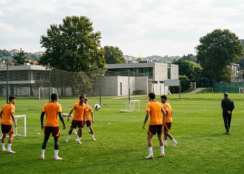 Futbolcular çim sahada antrenman yaparken antrenör onları izliyor.